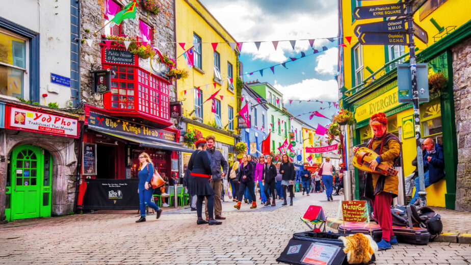 Vibrant street scene with colourful shopfronts, lively crowd, and street musician in charming Galway, Ireland.