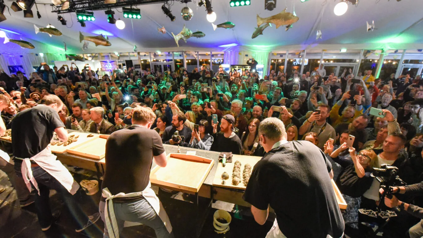 Three people shuck oysters at tables on stage as a large crowd watches and takes photos in a brightly lit auditorium decorated with hanging fish.