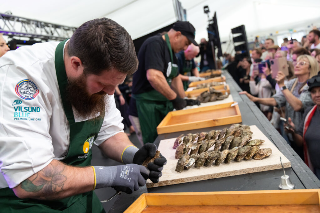 Three chefs shuck oysters at a competition, while a crowd of onlookers watches and takes photos in a large tented venue.