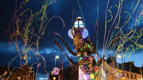 A large, colorful mechanical insect illuminated at night moves through a crowd, with people celebrating and colorful streamers flying through the air.