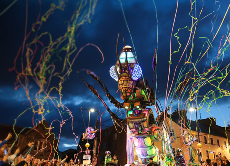 A large, colorful mechanical insect illuminated at night moves through a crowd, with people celebrating and colorful streamers flying through the air.
