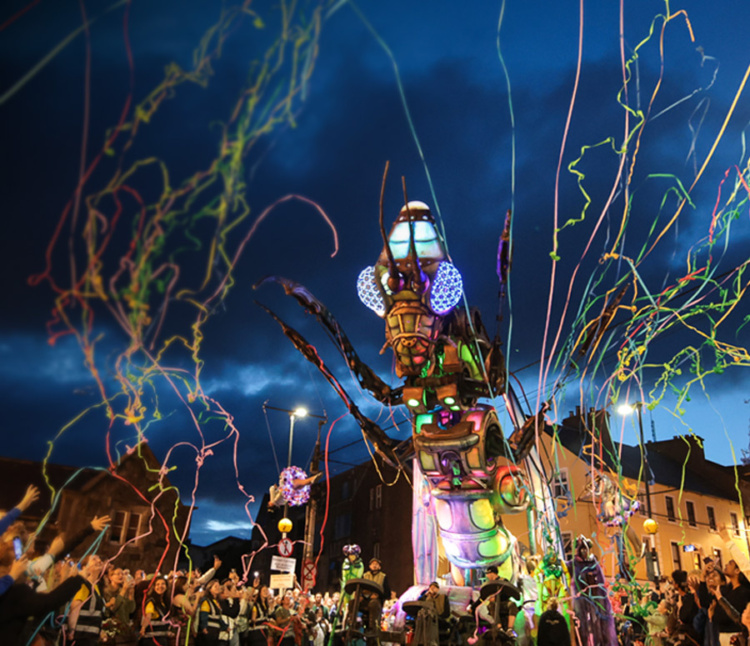A large, colorful mechanical insect illuminated at night moves through a crowd, with people celebrating and colorful streamers flying through the air.