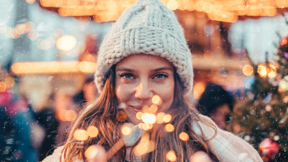Woman in knit hat enjoys sparkling festive lights at snowy Christmas market, creating a warm and joyful holiday atmosphere.