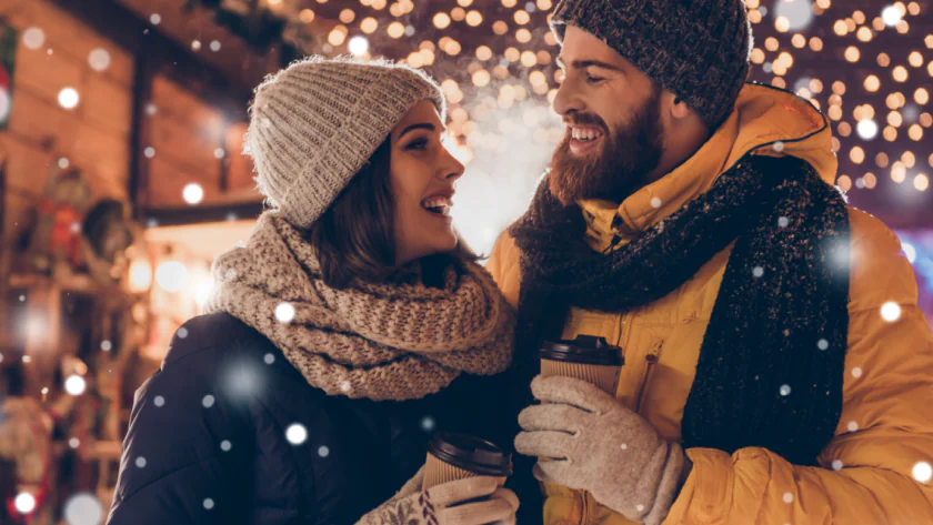Happy couple in warm clothes enjoying hot drinks in a snowy festive market under twinkling lights near cosy hotel.