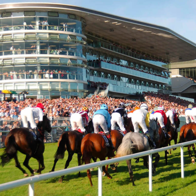 Jockeys riding horses around a bend during a race, with a packed grandstand and spectators in the background on a sunny day.