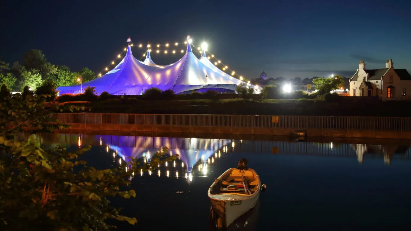 Illuminated marquee by a serene river, with cosy riverside cottages at night; perfect for romantic getaways.