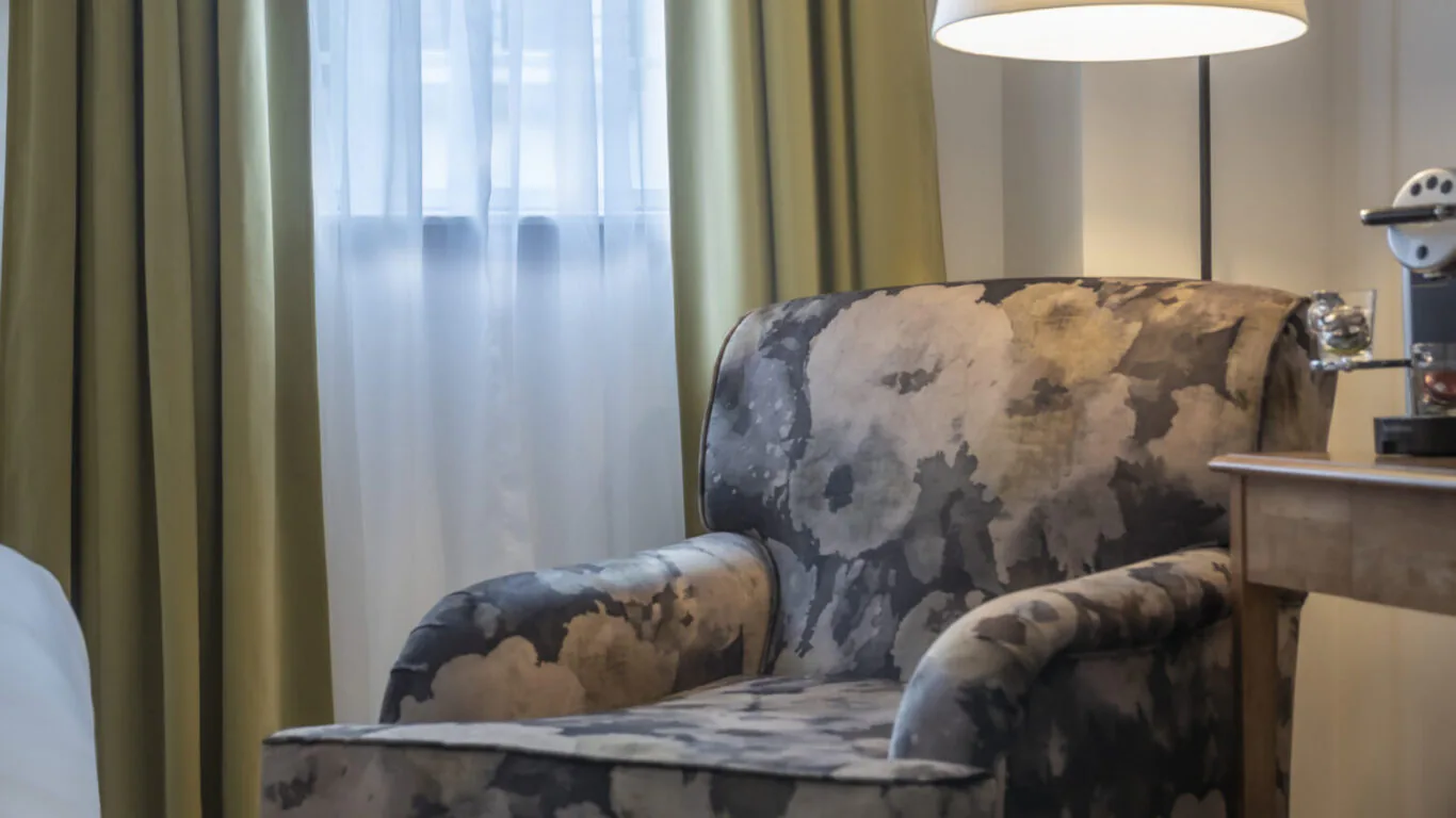 Cosy hotel room with floral armchair, warm lighting, and elegant green curtains, offering a relaxing atmosphere.