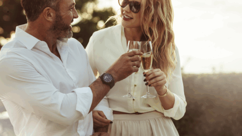 A man and woman in light clothing clink champagne glasses outdoors, smiling at each other in a sunlit setting.