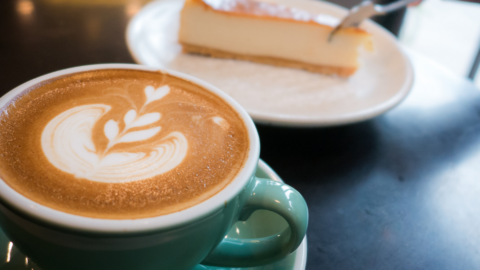 A cup of latte with leaf latte art in a green mug sits on a table next to a plate with a slice of cheesecake and a fork.