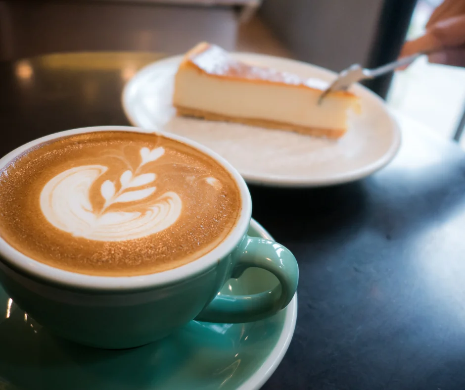 A cup of latte with leaf latte art in a green mug sits on a table next to a plate with a slice of cheesecake and a fork.
