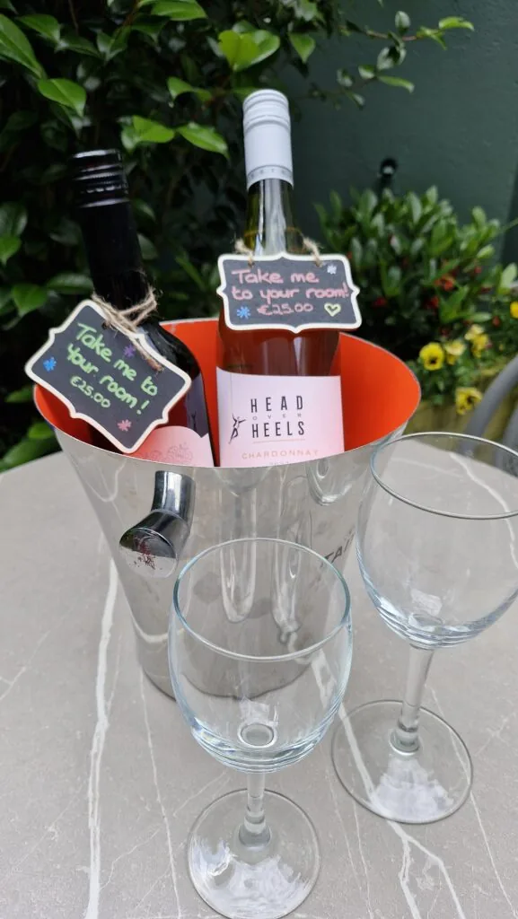 Two bottles of wine in an ice bucket with price tags, next to two empty wine glasses on an outdoor table with green plants in the background.