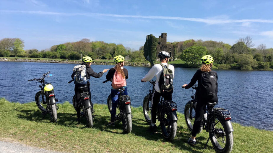 Four people wearing helmets and backpacks stand with their bikes by a lake, facing the water and a distant castle under a clear blue sky.