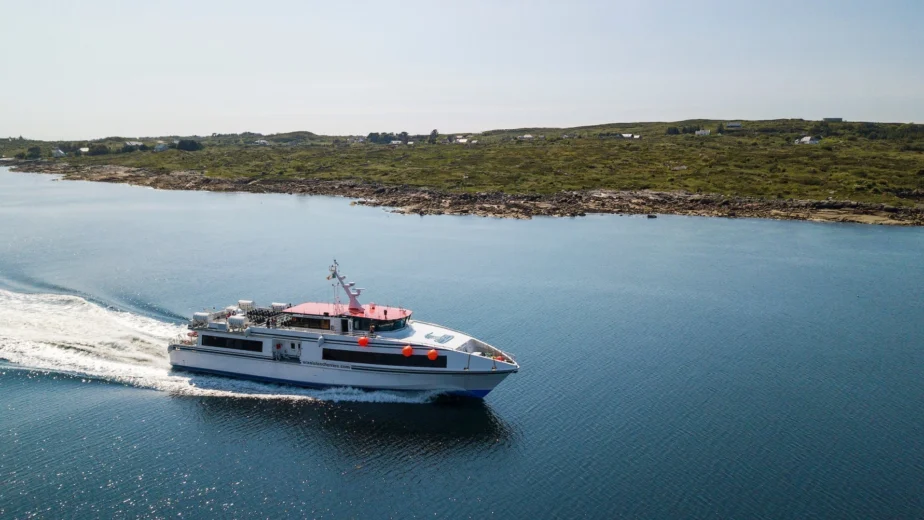 A passenger ferry travels across calm water near a rocky shoreline with scattered buildings under a clear sky.