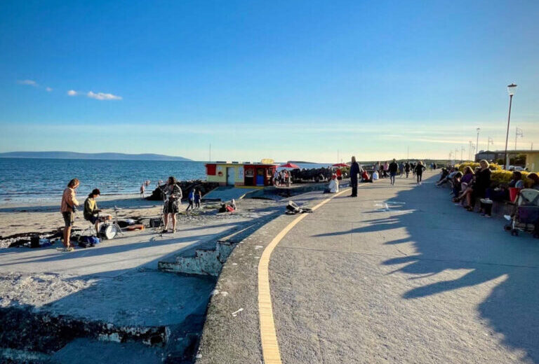 People gather along a seaside promenade on a sunny day, with some sitting, walking, and standing near the water. The sky is clear and the sea is calm.