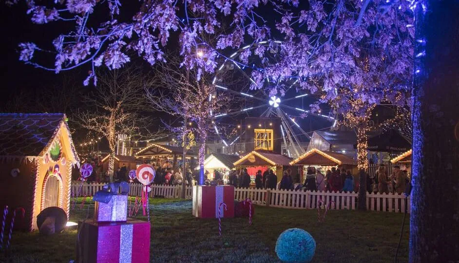 Nighttime scene of a festive holiday fair with decorated trees, gingerbread house displays, wrapped gifts, and a lit Ferris wheel in the background. Crowds are visible behind a white fence.