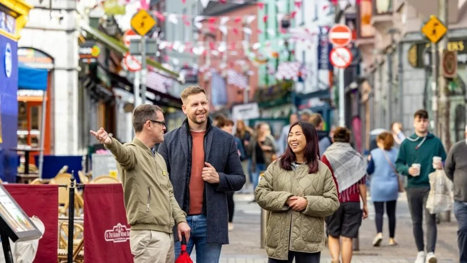 Three people walk and talk on a lively, colorful street decorated with bunting; other pedestrians and shopfronts are visible in the background.