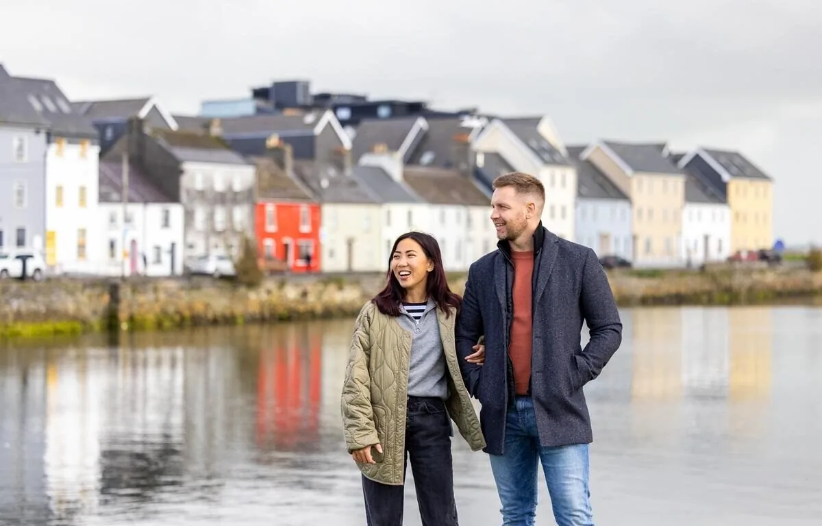 A man and woman stand together by a waterfront with colorful houses in the background on a cloudy day.