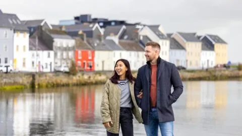 A man and woman stand together by a waterfront with colorful houses in the background on a cloudy day.