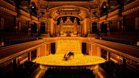 A grand concert hall with ornate architecture, featuring a pianist performing onstage surrounded by numerous glowing candles.