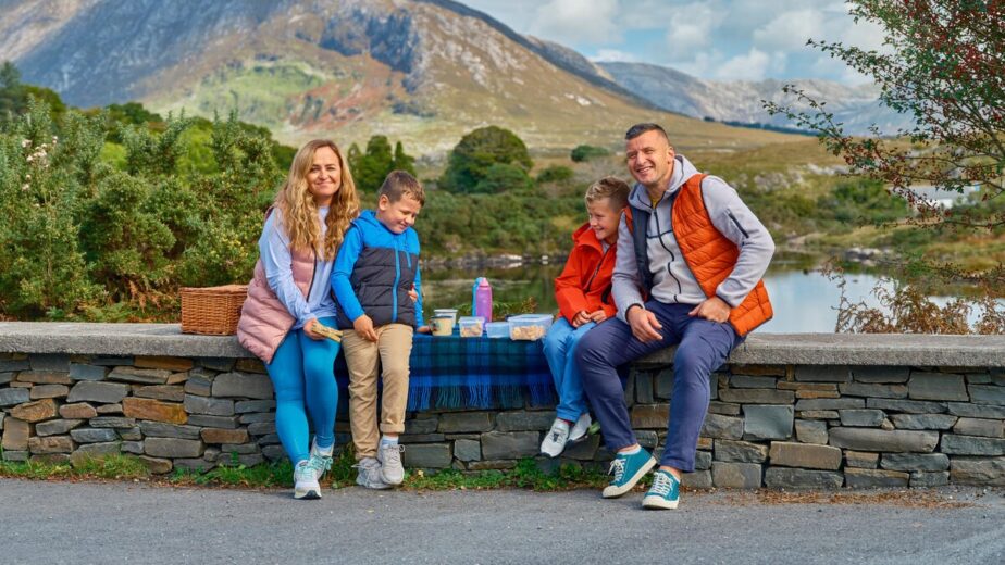 A family of four sits on a stone wall having a picnic with food and drinks, with mountains, trees, and a lake in the background.
