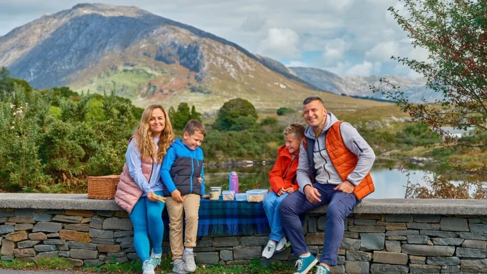 A family of four sits on a stone wall having a picnic with food and drinks, with mountains, trees, and a lake in the background.