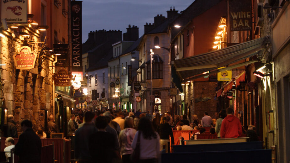 Lively evening street with guests enjoying al fresco dining, surrounded by glowing signs and charming stone architecture.
