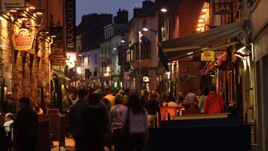 Lively evening street with guests enjoying al fresco dining, surrounded by glowing signs and charming stone architecture.